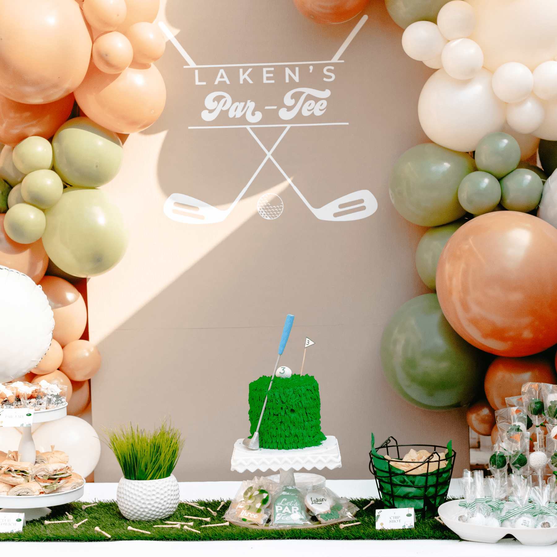 A picture of the birthday cake on a table with other snacks and balloons in the background.
