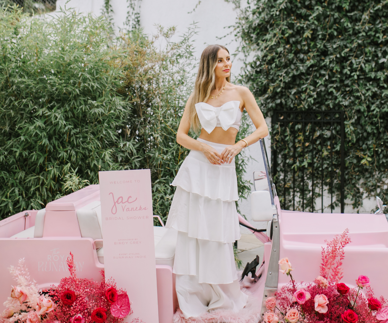 A woman in a white dress standing on a pink, vintage car.