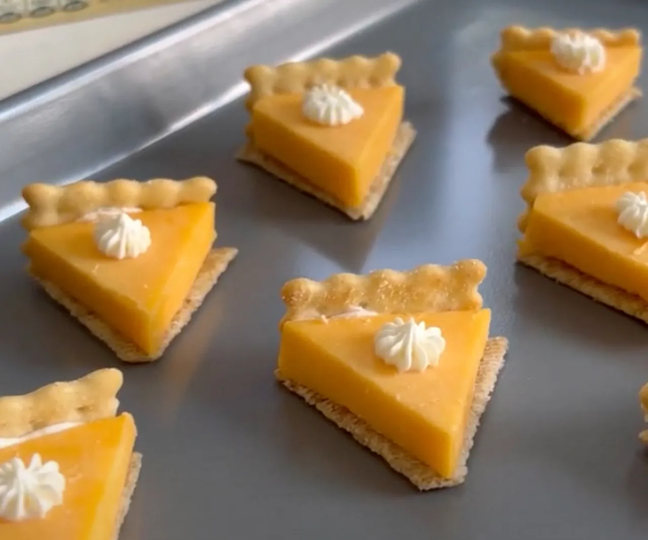 Small triangular orange cheese slices on square crackers topped with small white dollops on a metal tray.