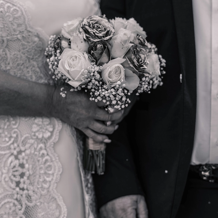Black and white photo of a close up of a bride's bouquet, standing next to a man in a tux