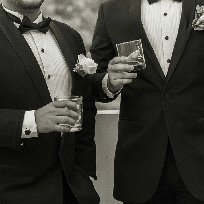 Black and white photo close up of wedding formal attire, featuring two men in tuxedos holding whiskey glasses.