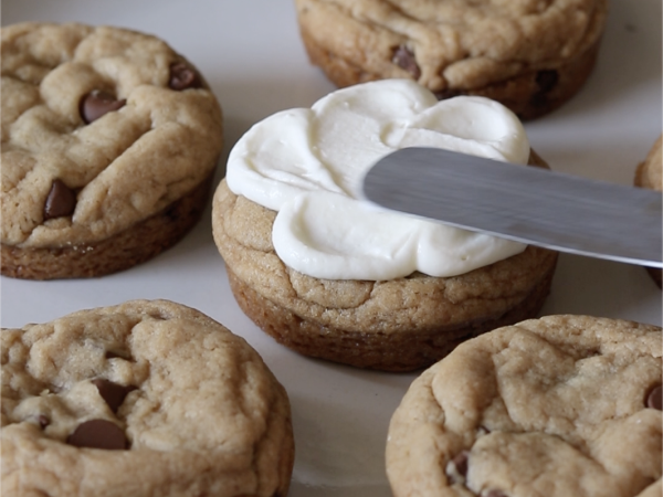 Close up of frosting being spread over a chocolate chip cookie.