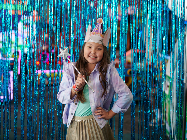 A girl in a Chuck E. Cheese birthday hat holding a star wand in front of a blue tinsel backdrop.