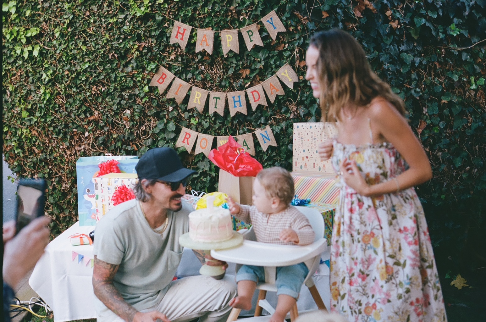 Parents standing next to the birthday boy as they bring him his birthday cake.