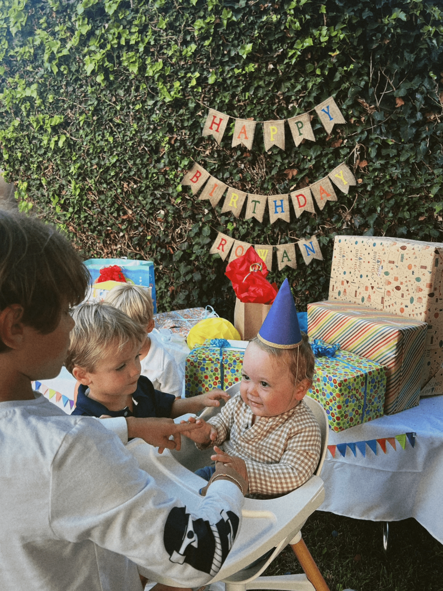 Kids gathering around the birthday boy in his high chair with a table full of presents in the background.
