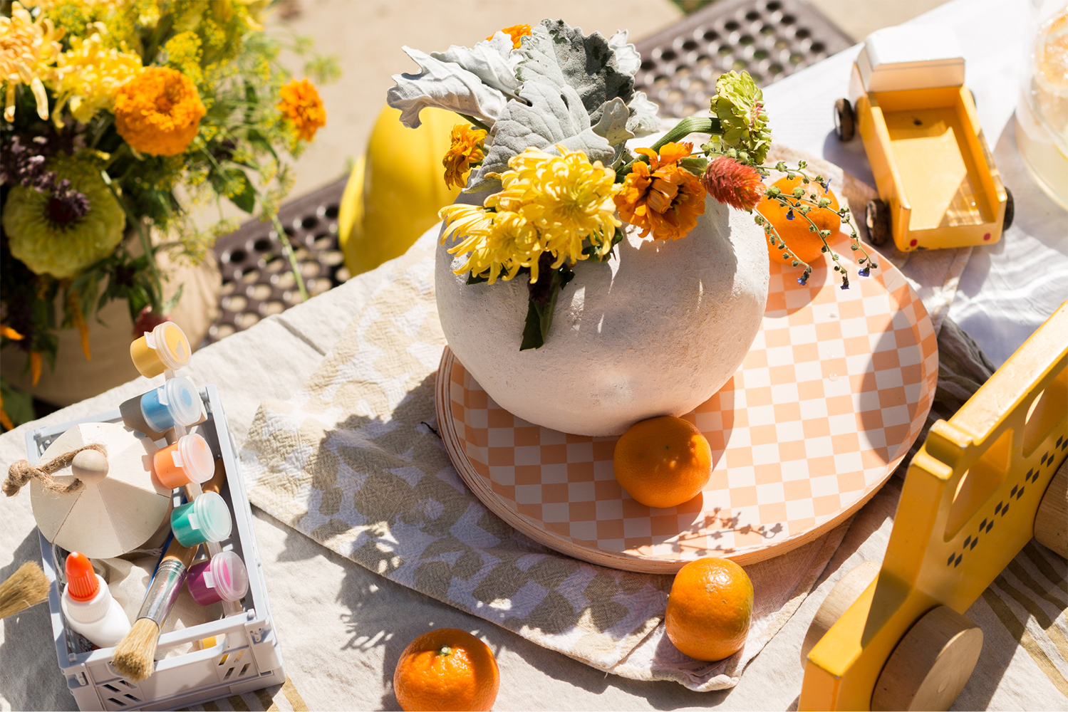 A table with paint and a vase full of orange and yellow flowers.