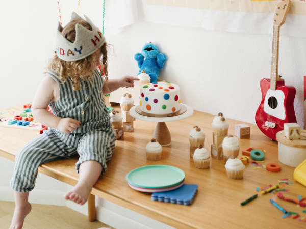 Child siting on a table next to a cake, cupcakes and toys.